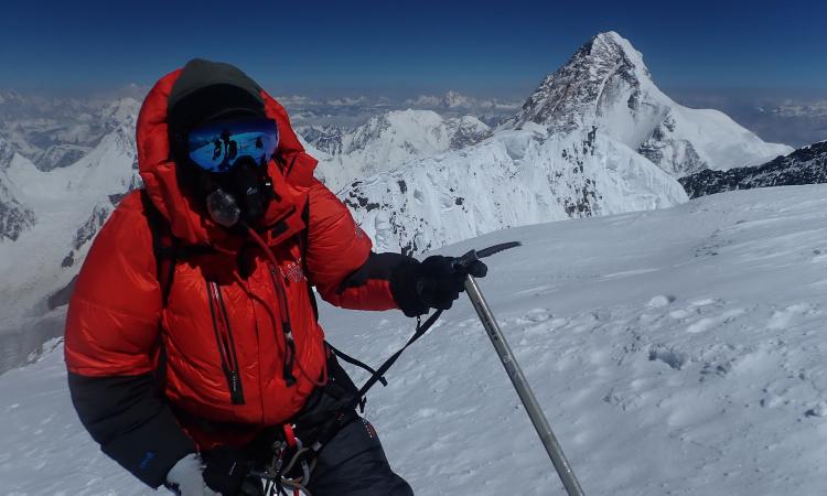 Matthew Holt summiting Broad Peak in heavy snow, with K2’s towering peak visible in the background, capturing the extreme conditions of high-altitude mountaineering in the Karakoram Range.